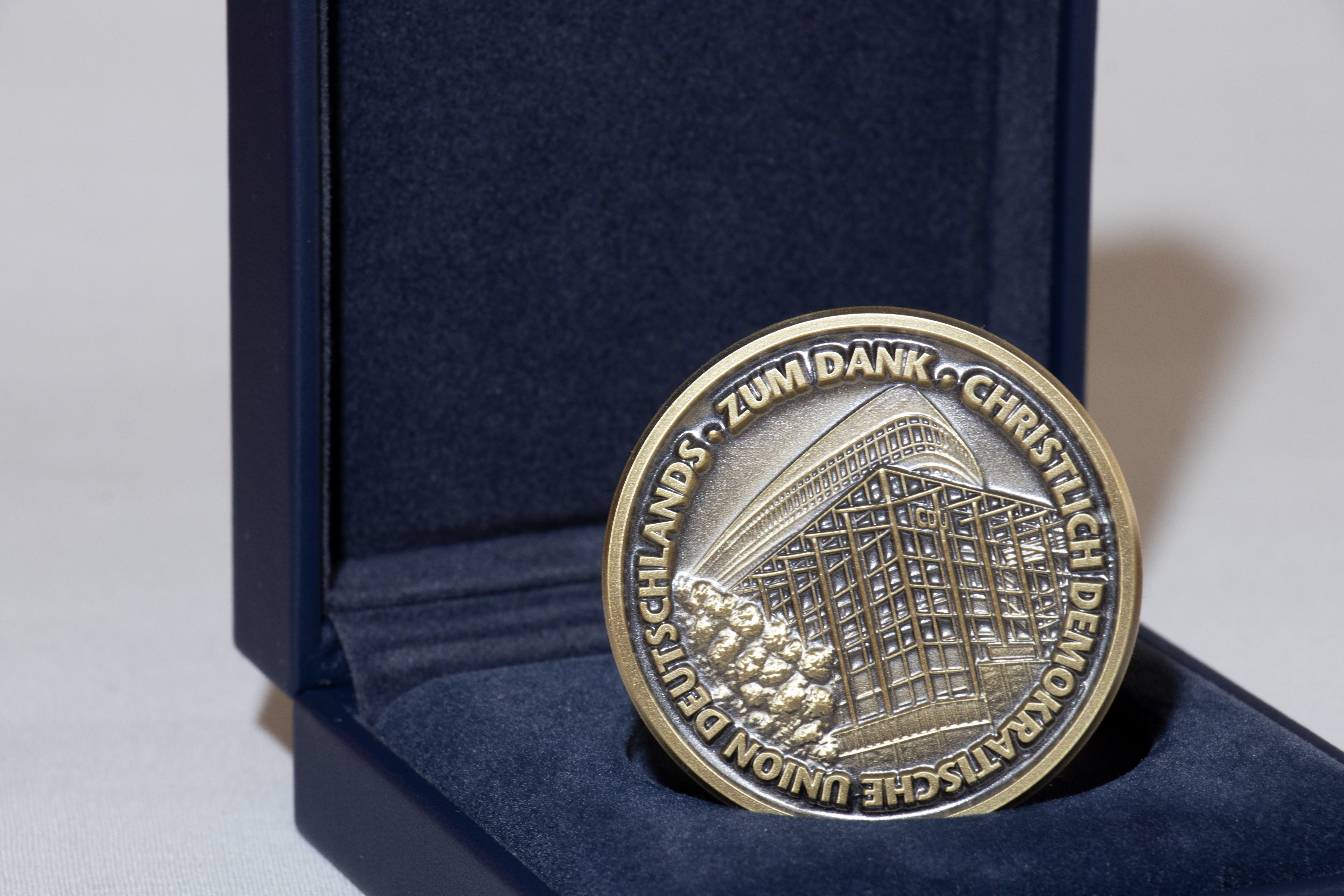 Bildausschnitt größer: Aufgeklappte Schachtel mit ausgelegtem Samt in Dunkelblau. Darin aufrecht gesteckt eine Medaille in Bronze. Mit Prägung: Zum Dank Christlich Demokratischer Union Deutschlands